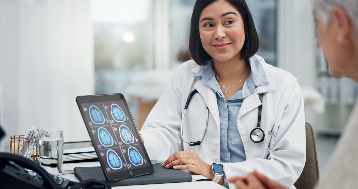 A doctor at a desk with a tablet showing brain scans, talking with a person across the desk