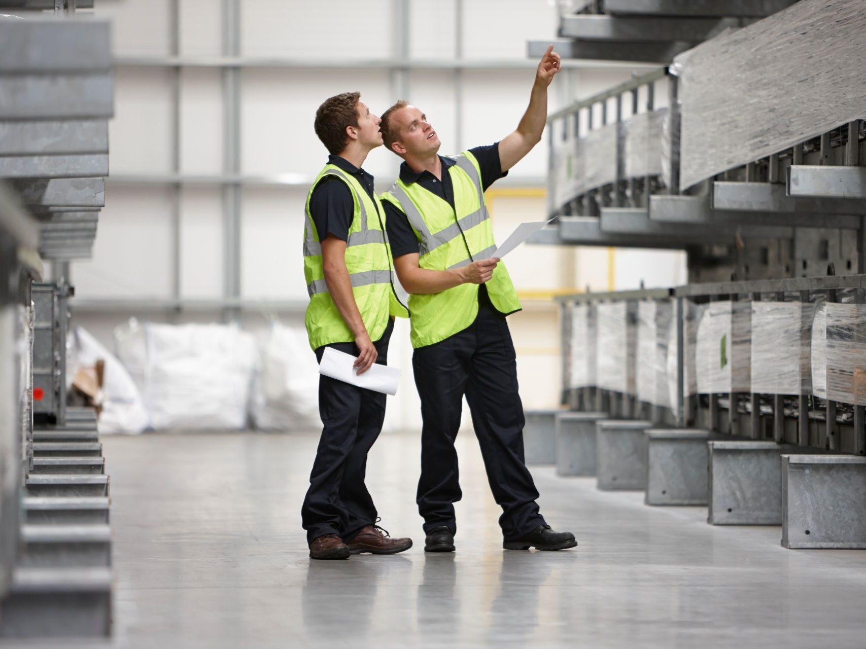 Two warehouse workers in high-visibility vests checking stock on tall racks against a printed list