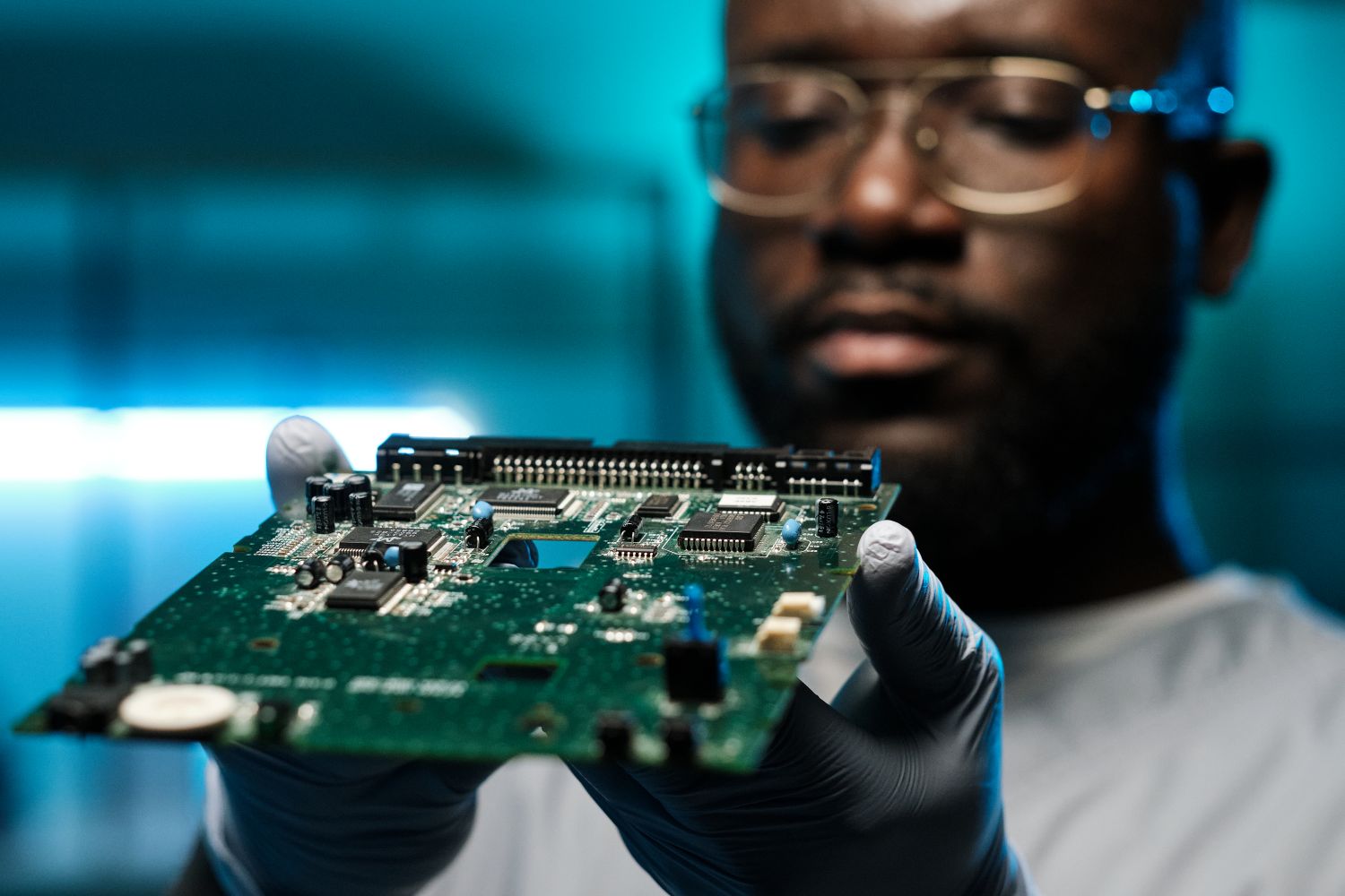 An engineer in glasses and dark gloves holding up a printed circuit board to inspect it