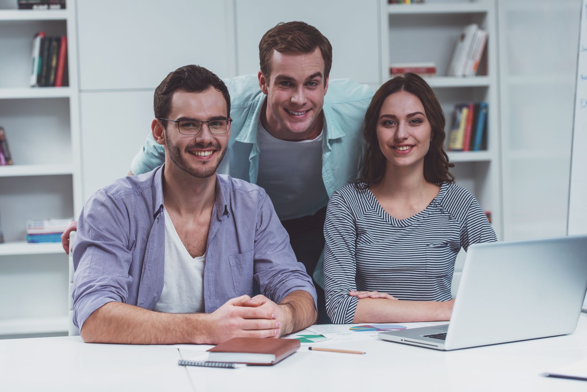 Three smiling colleagues gathered at a desk around a laptop in a bright office