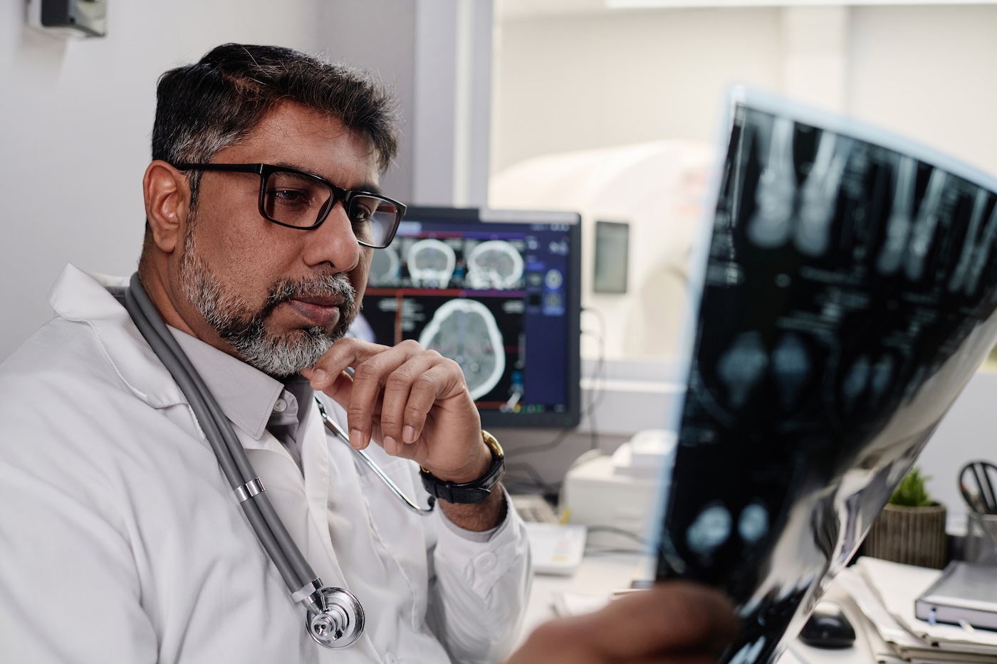 A doctor in glasses studying an X-ray film, with scan images on the monitor behind him