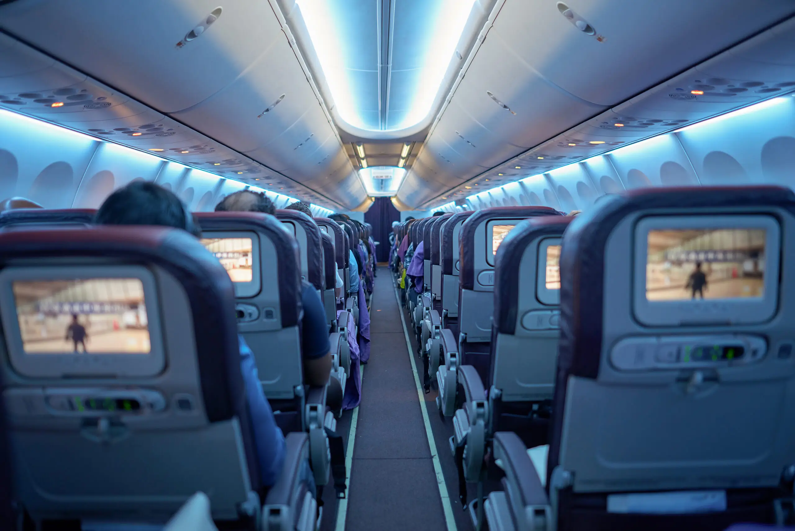 The aisle of an aircraft cabin between rows of seats with seat-back screens, passengers seen from behind