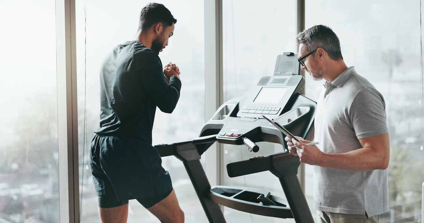 A man running on a treadmill by a tall window while a trainer beside him checks a tablet