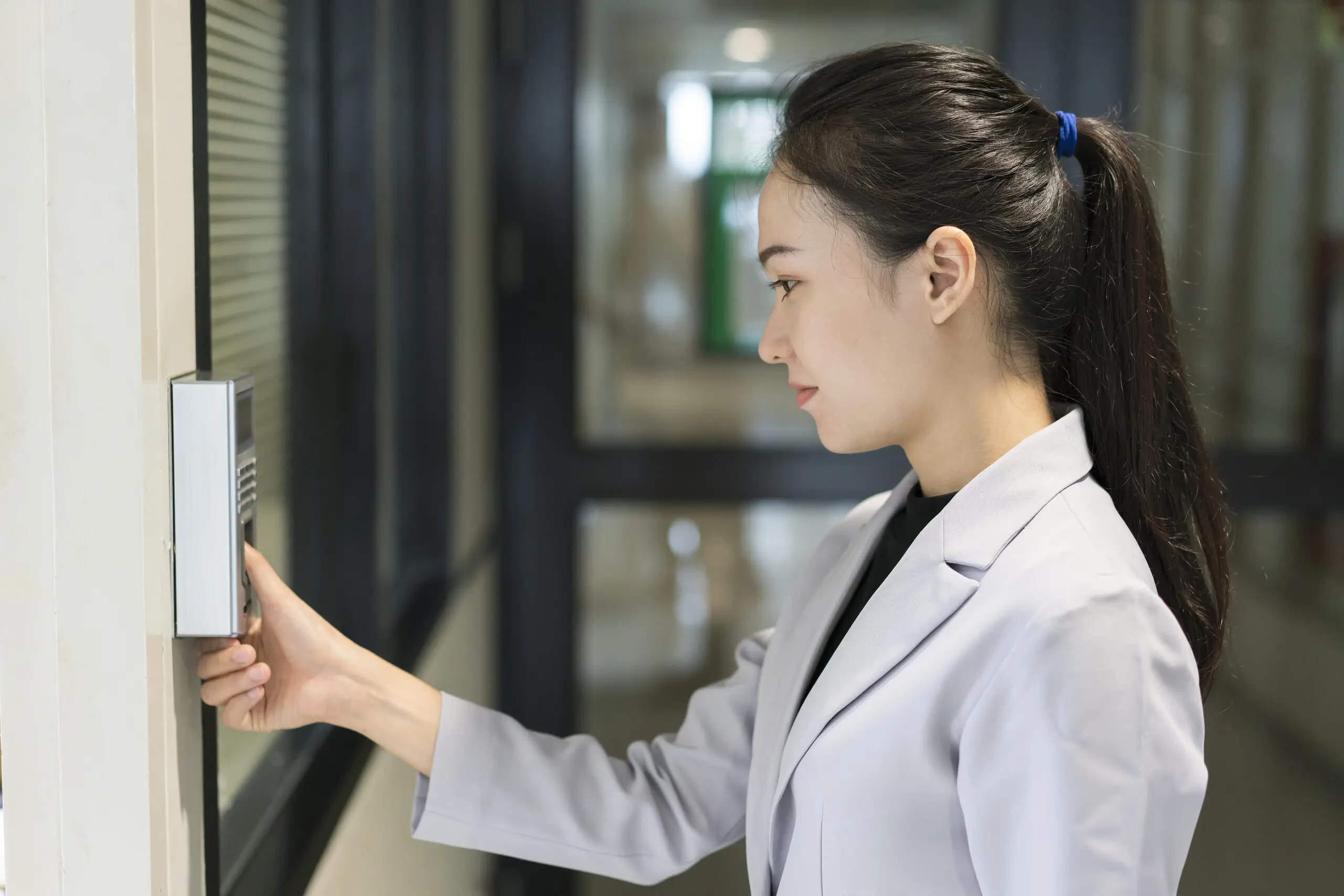 A woman in a light jacket pressing her finger to a wall-mounted access reader in a corridor