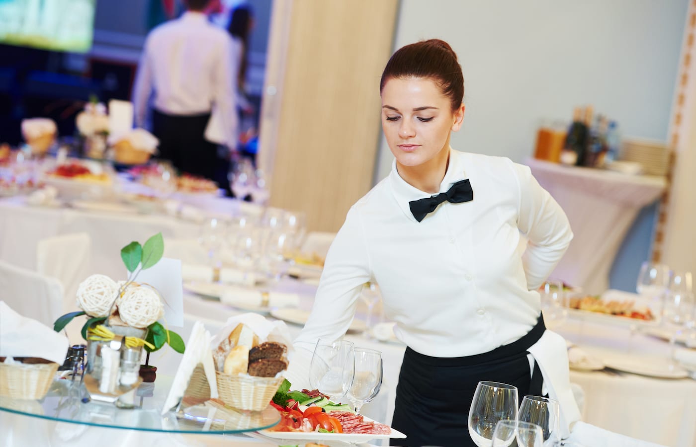 A waitress in a white shirt and bow tie setting a banquet table laid with glasses and food