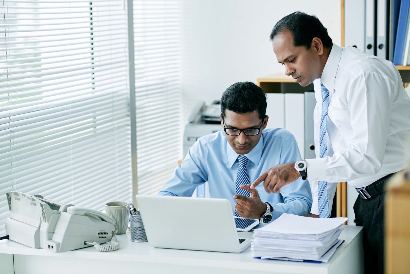 A manager pointing at a laptop screen for a colleague at a desk stacked with paper files