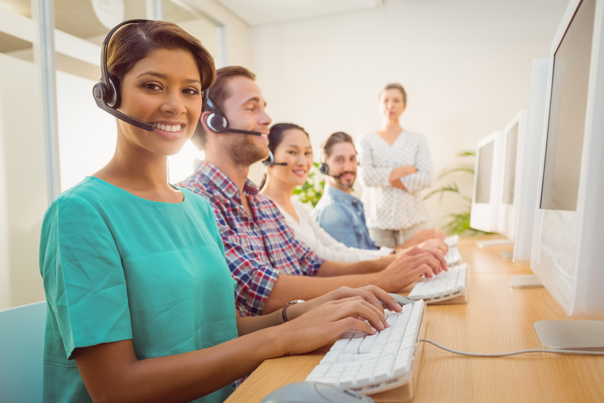 A row of customer-service agents in headsets typing at computers, with a supervisor standing behind them