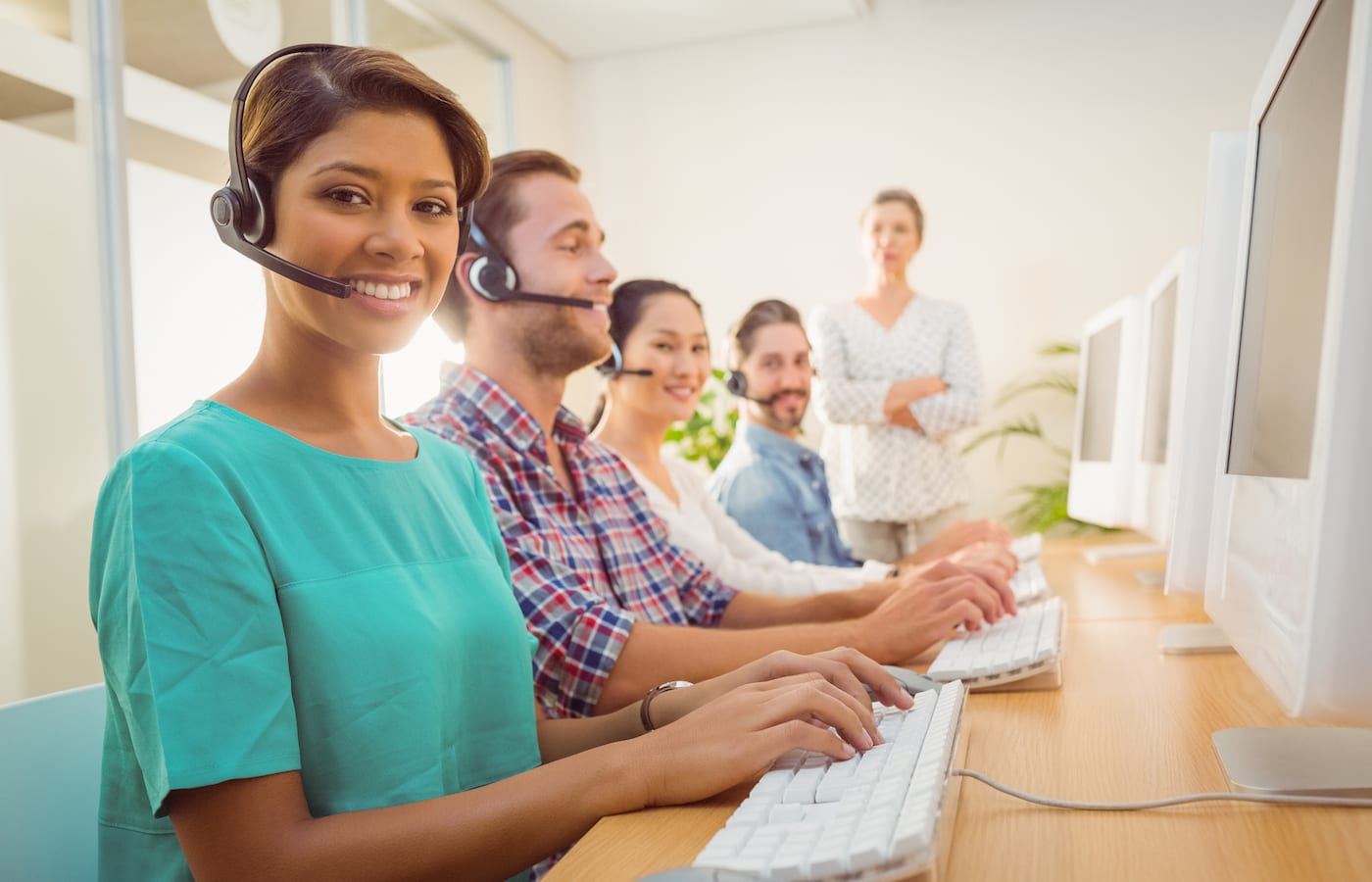 A smiling agent in a headset at a computer, with a row of colleagues in headsets behind her