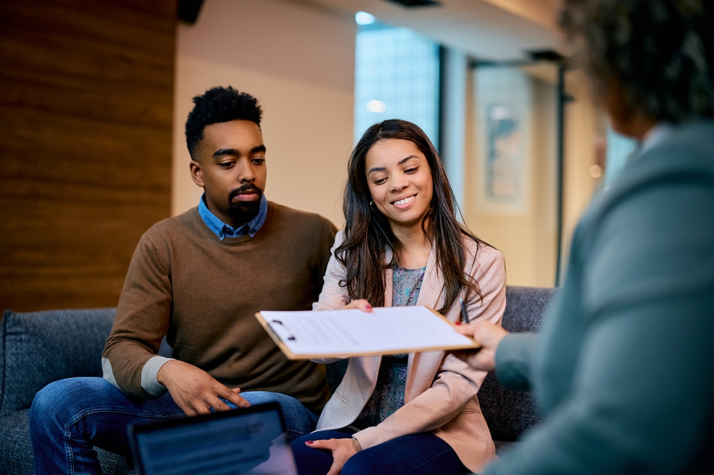A couple on a sofa at home reviewing a document on a clipboard with a visiting adviser