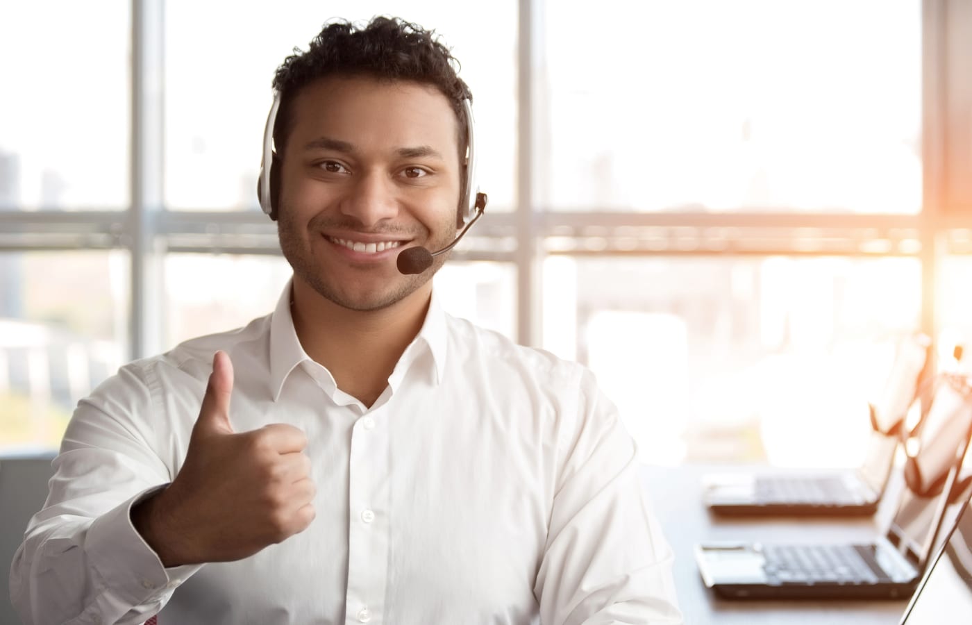 A smiling agent in a headset giving a thumbs-up at a desk by a bright window