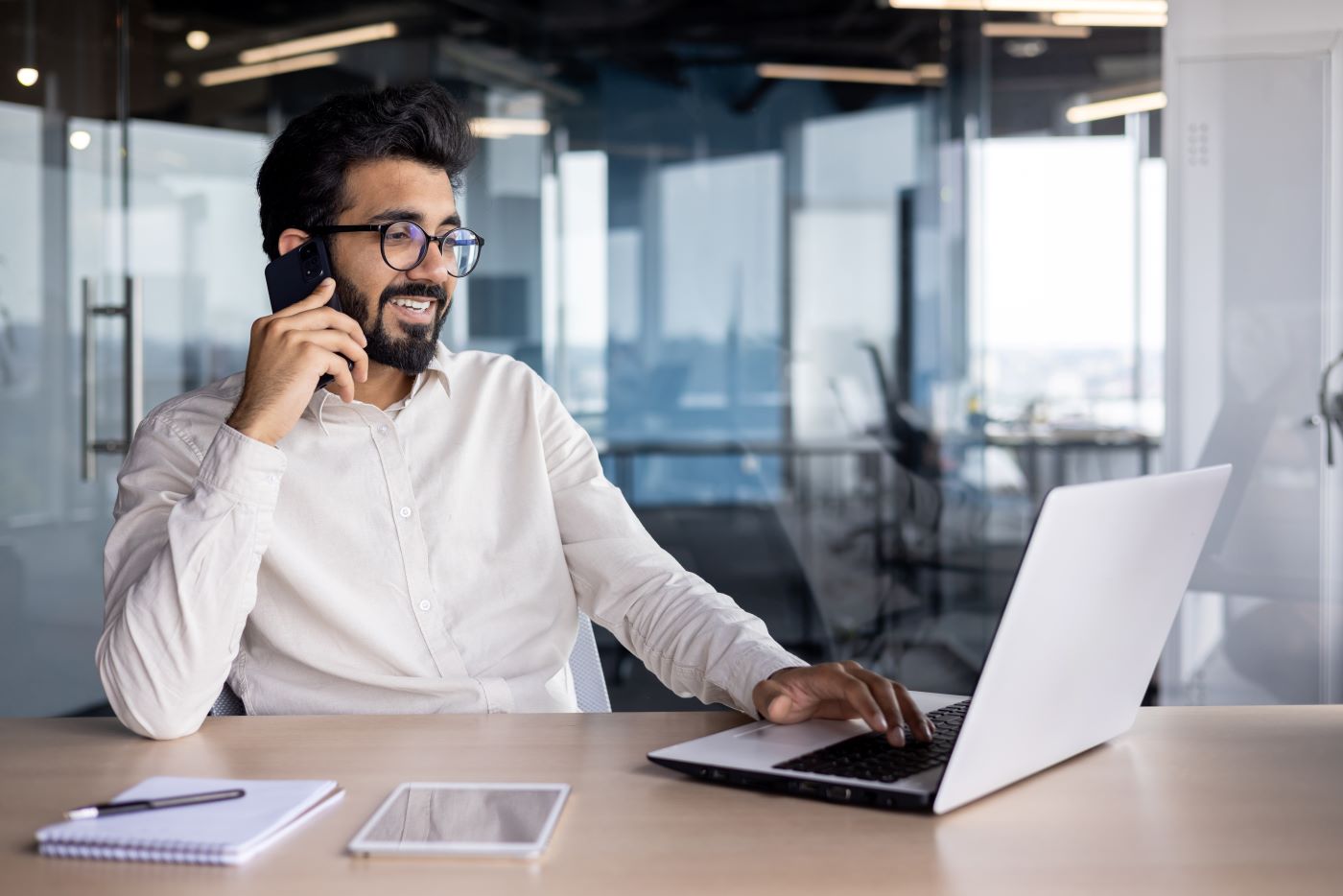 A man on a phone call at a laptop in a glass-walled office