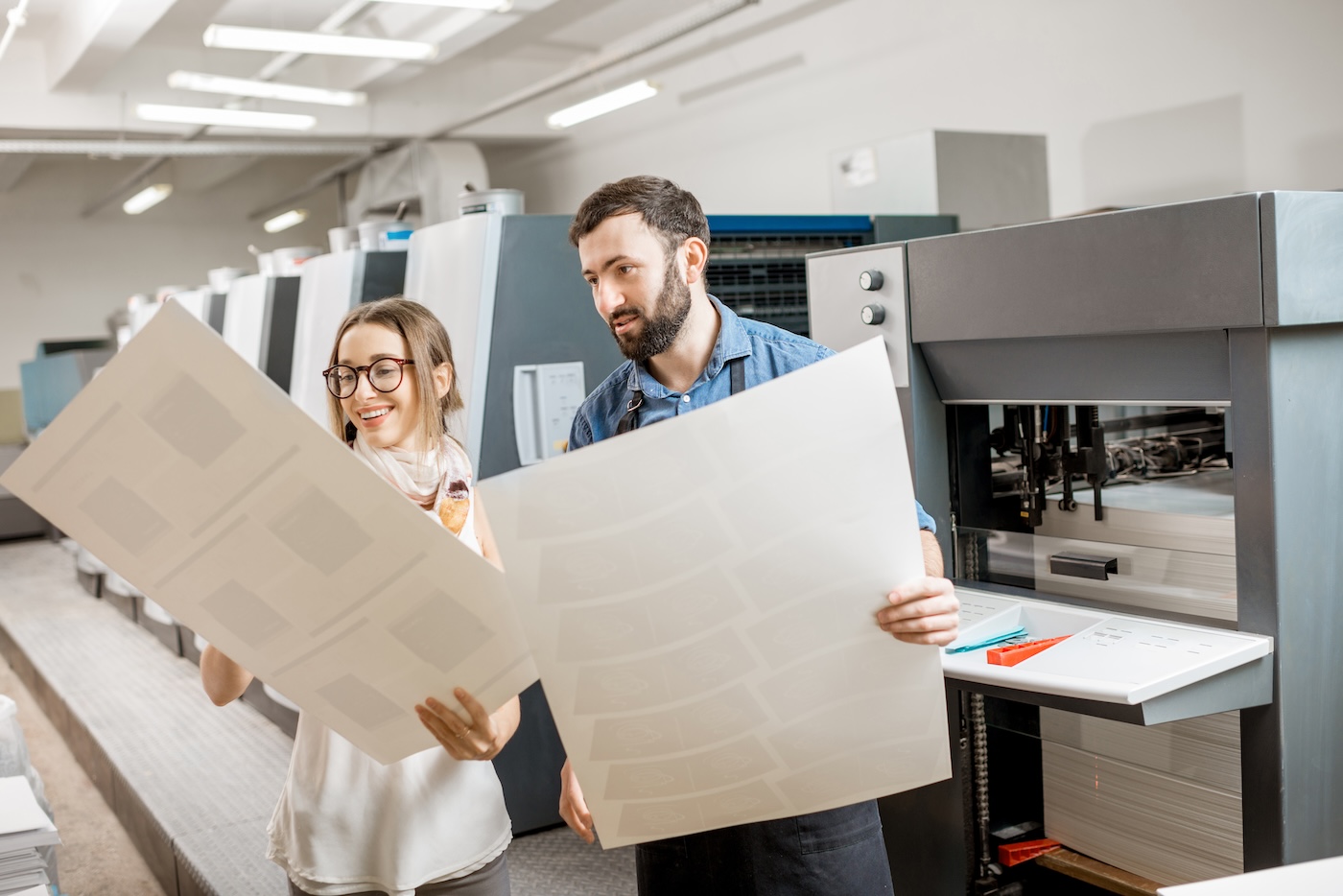 Two colleagues checking large printed proofs beside a printing press