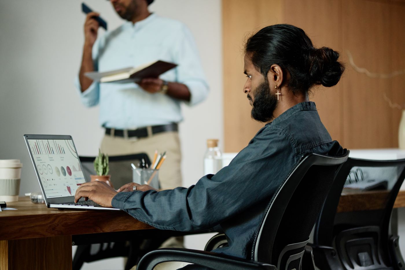 A man working on a laptop showing charts while a colleague reads notes behind him