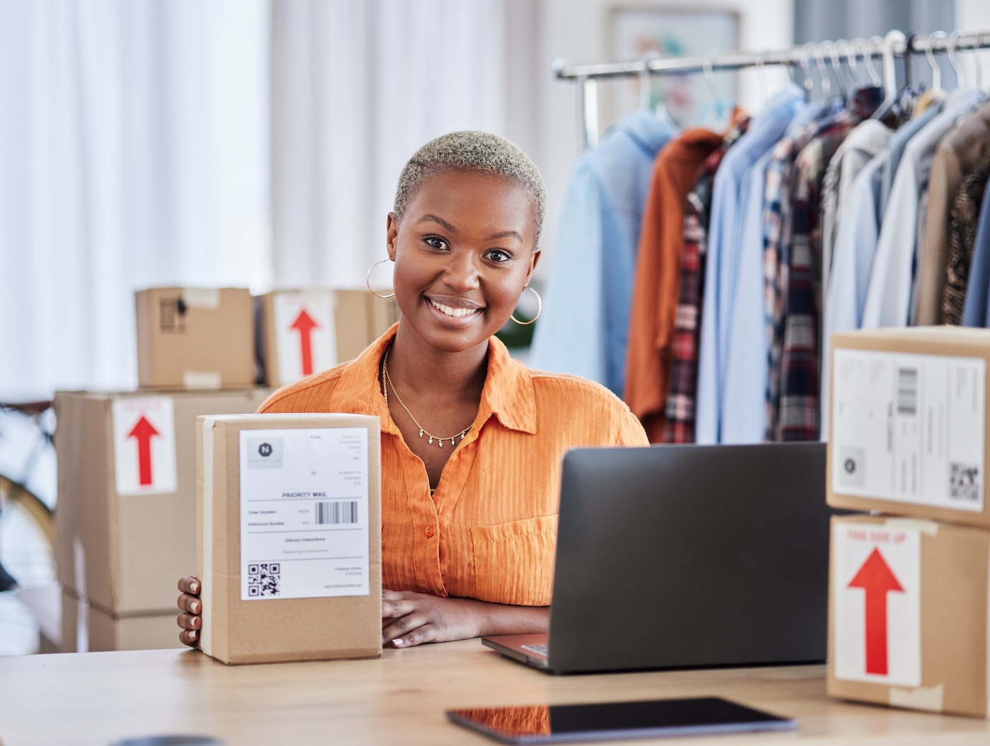 An online seller holding a labelled parcel beside a laptop and a rail of clothes
