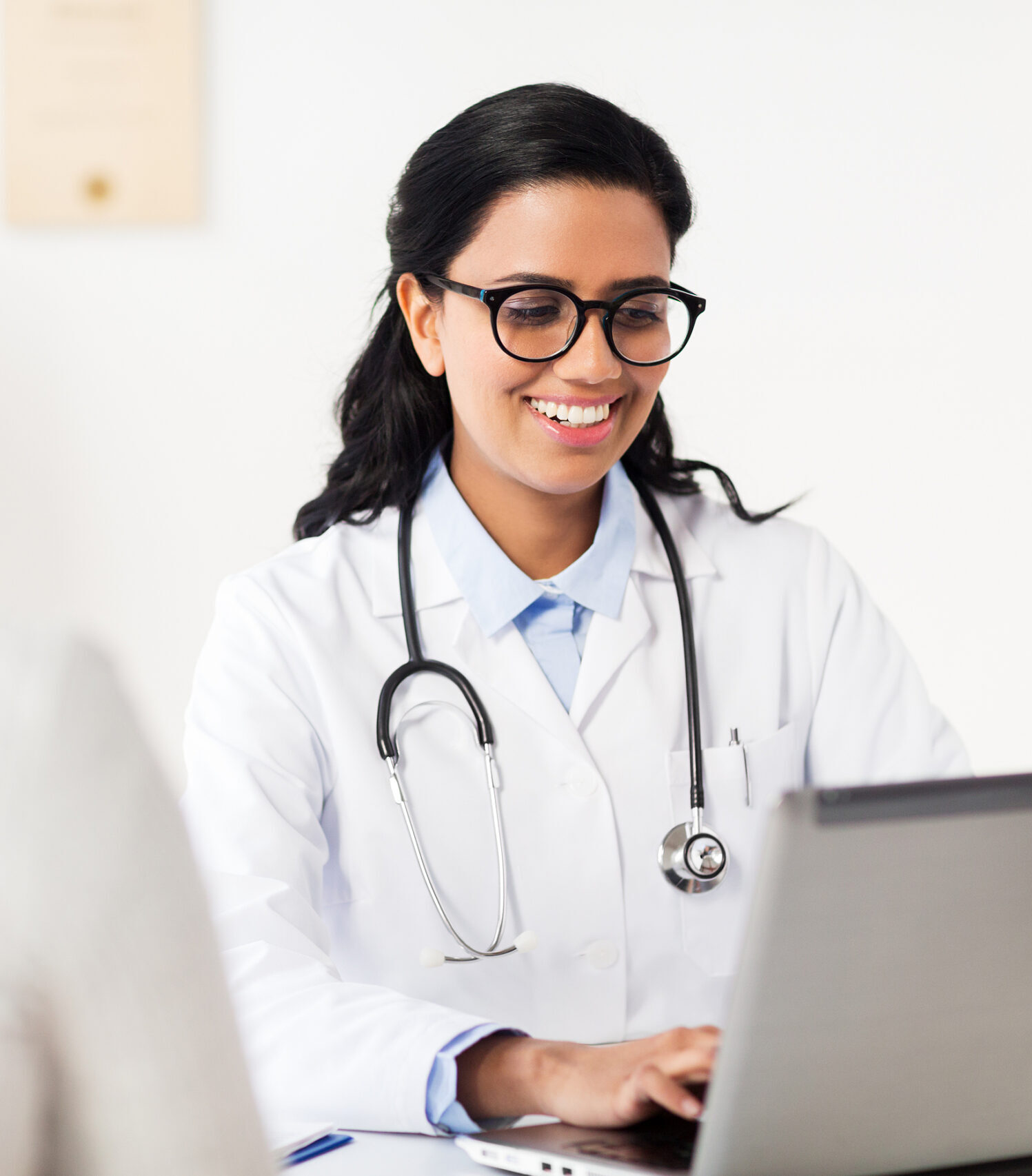 A smiling doctor in glasses with a stethoscope working on a laptop