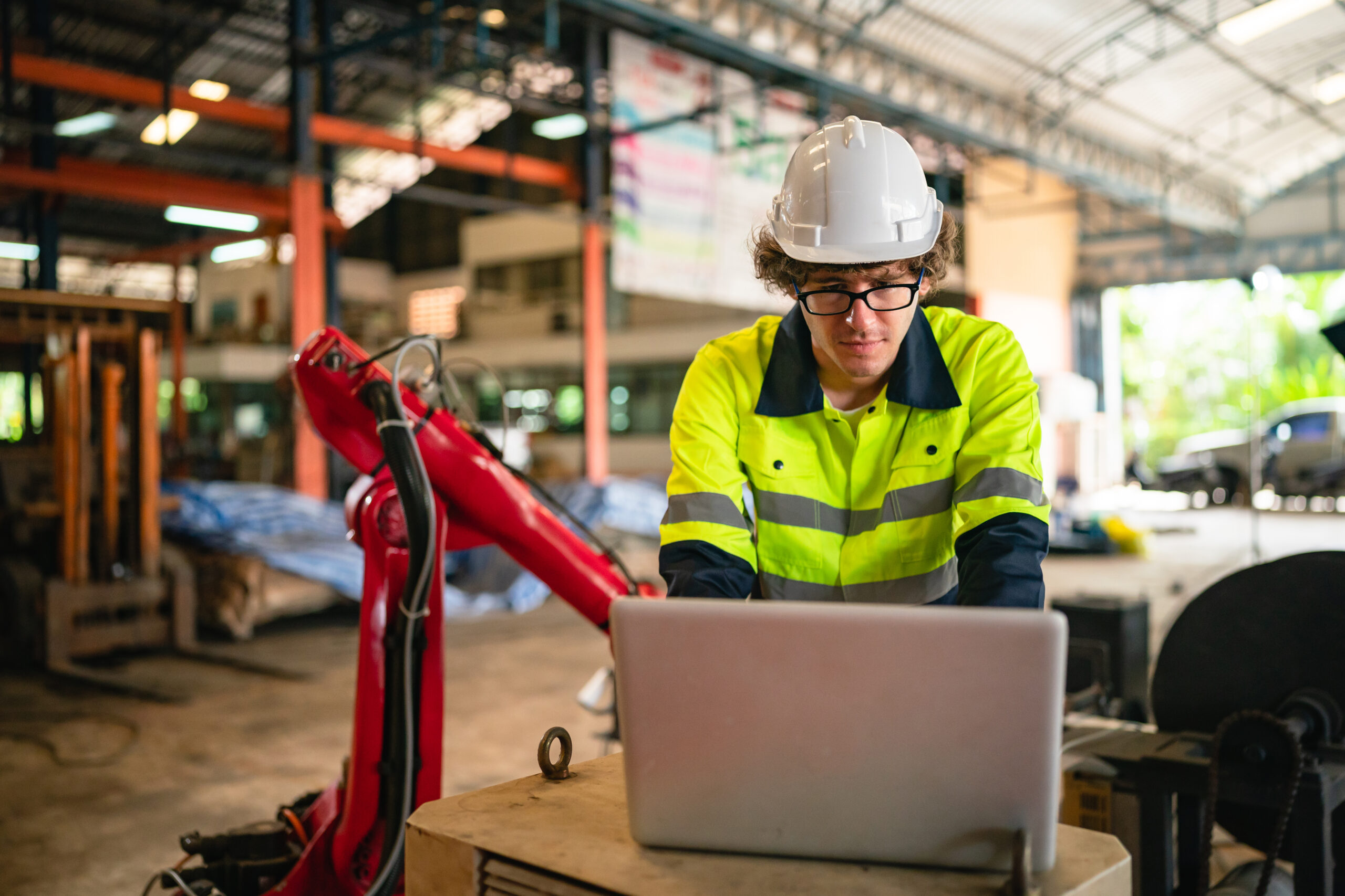 An engineer in a hard hat working on a laptop beside a robotic arm