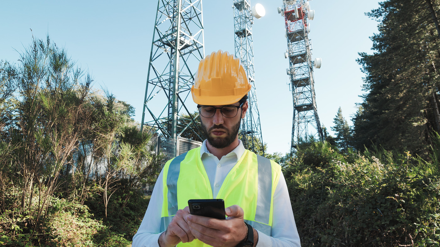 An engineer in a hard hat and high-visibility vest checking his phone below telecom masts