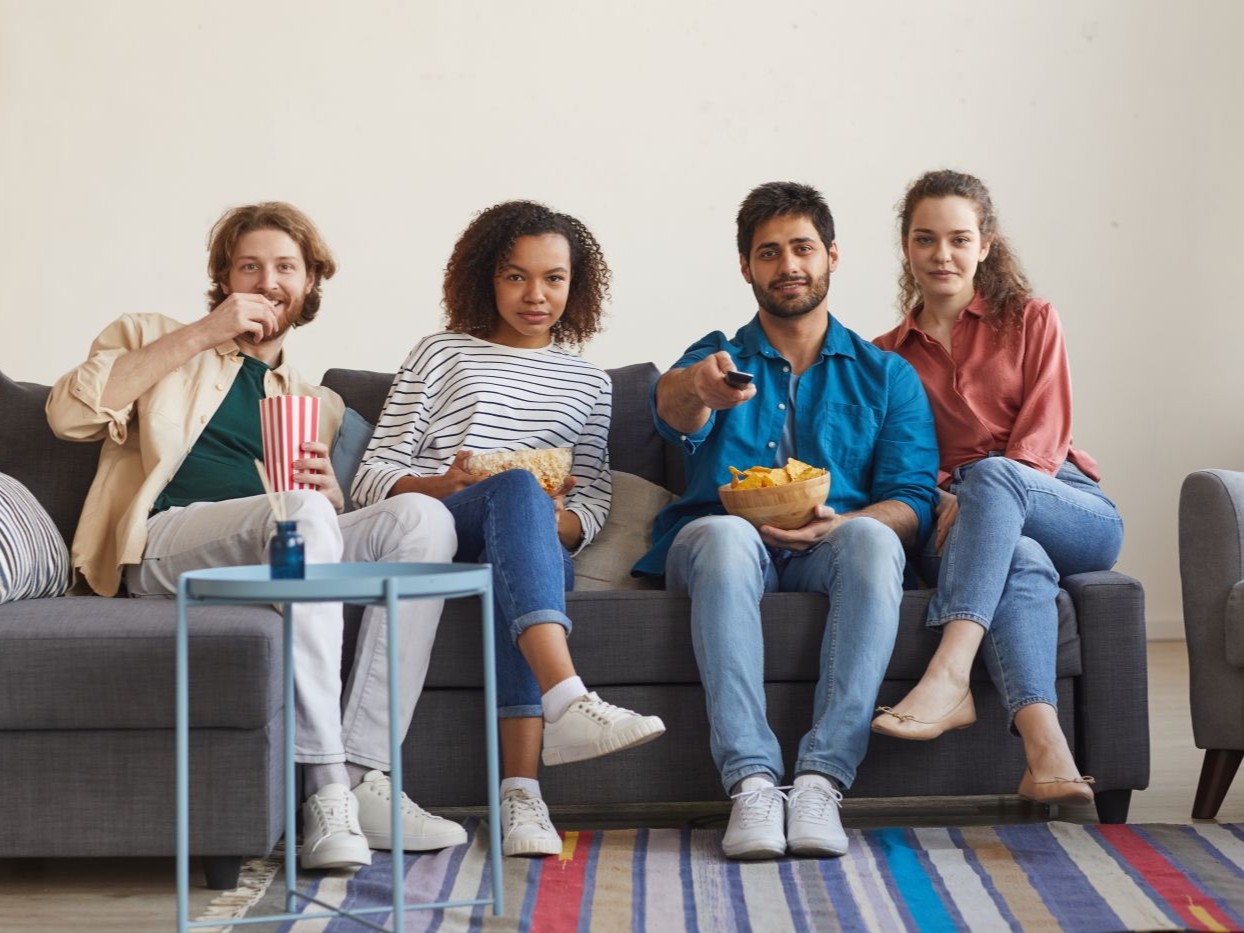 Four friends sitting on a sofa with popcorn, watching a screen together at home