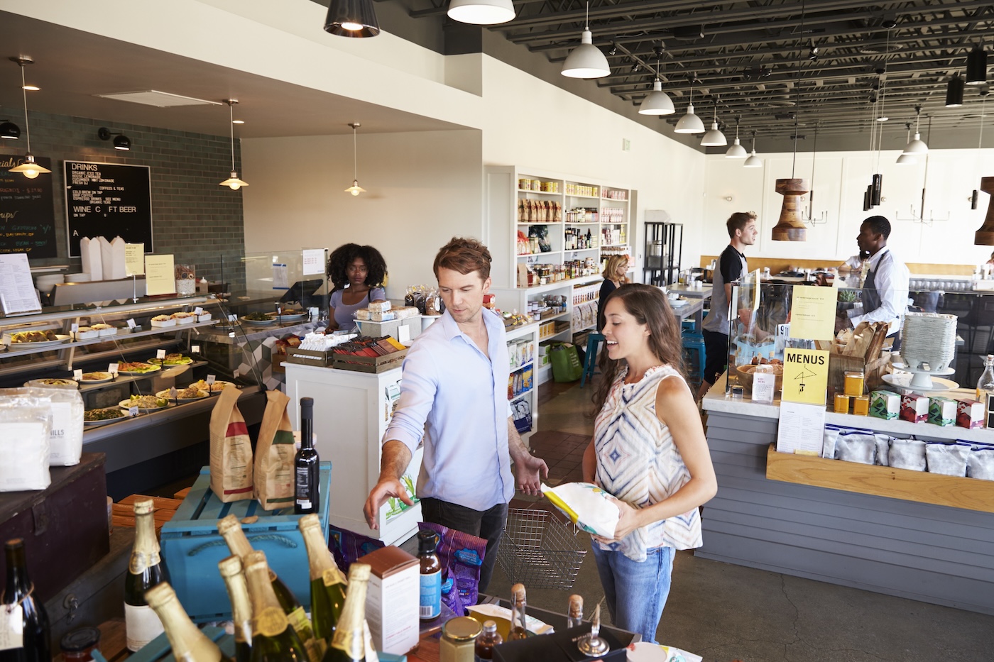 Shoppers with a basket browsing a delicatessen and grocery store while staff work behind the counters