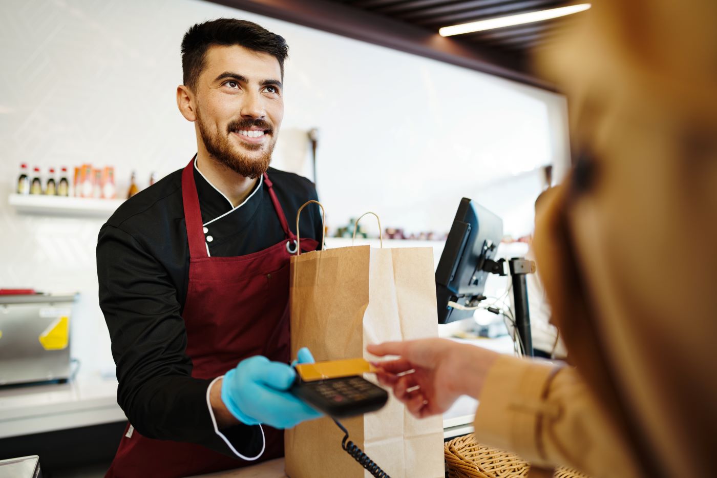 A shop assistant in gloves holding out a card terminal as a customer pays at the counter, a paper bag beside it