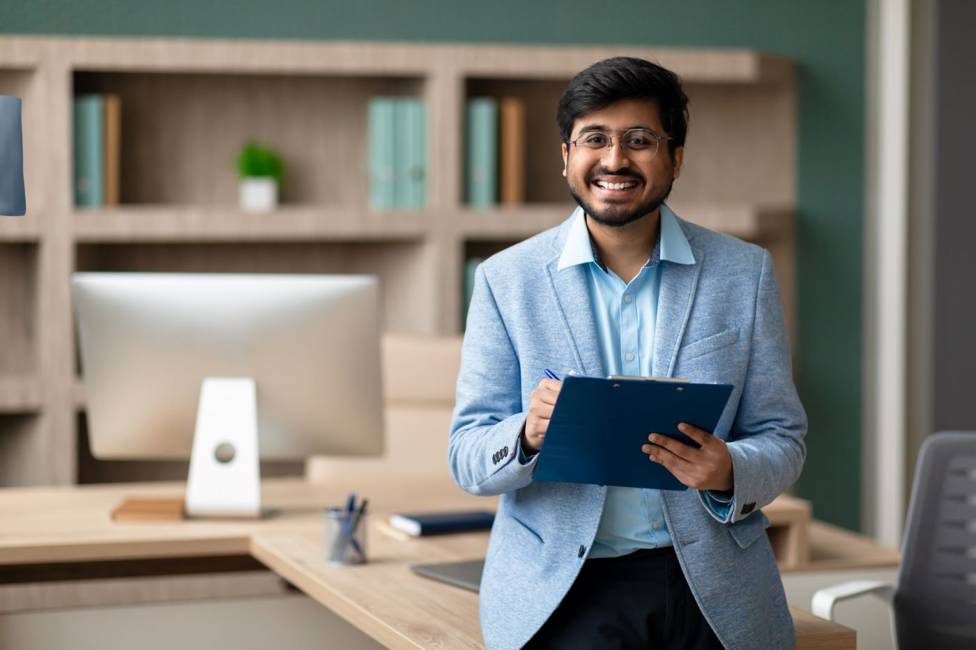 A smiling man in a light blazer making notes on a clipboard beside an office desk