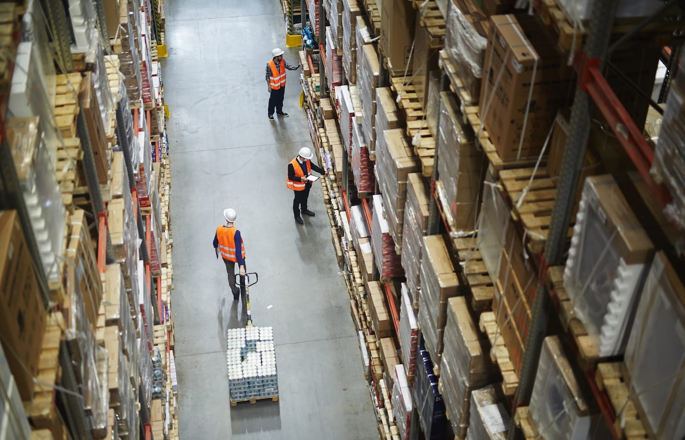 Workers in high-visibility vests in a tall warehouse aisle stacked with goods