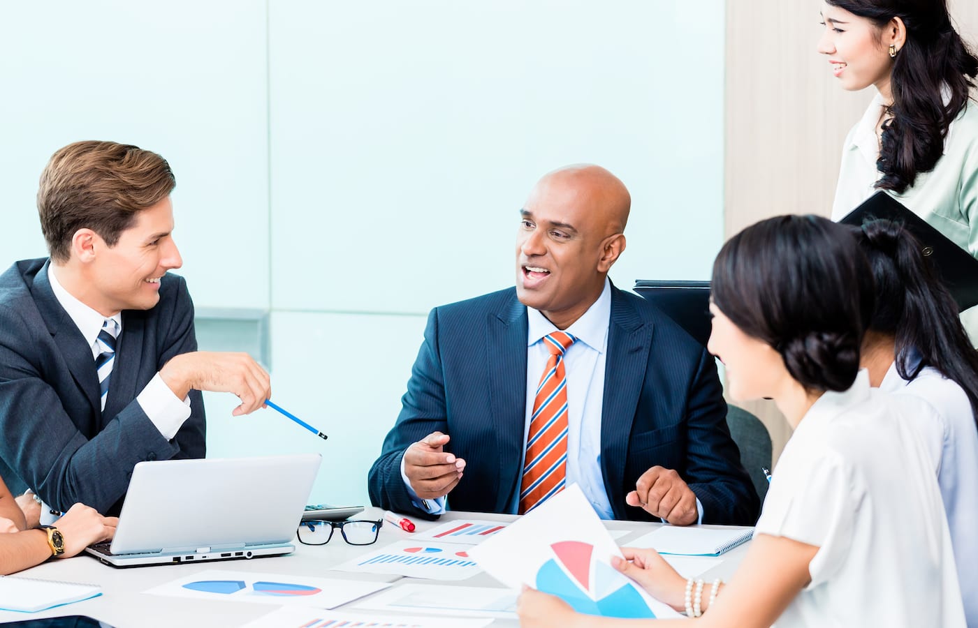 Colleagues in business dress talking around a meeting table with a laptop and printed charts