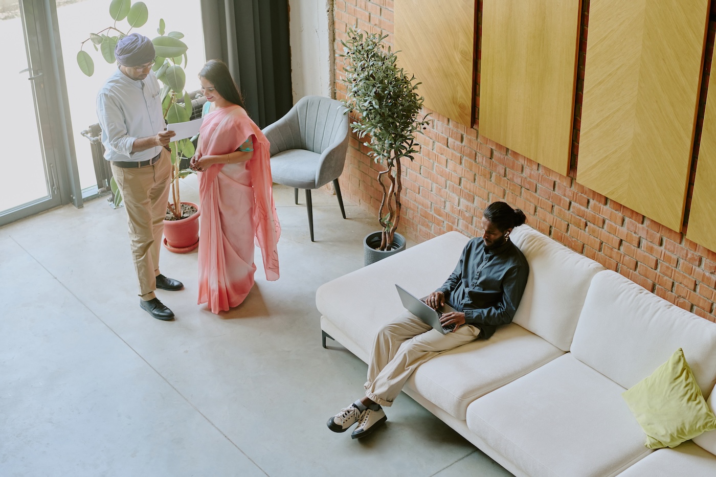 A lounge seen from above, a man working on a laptop on a sofa while two people talk beside the armchairs