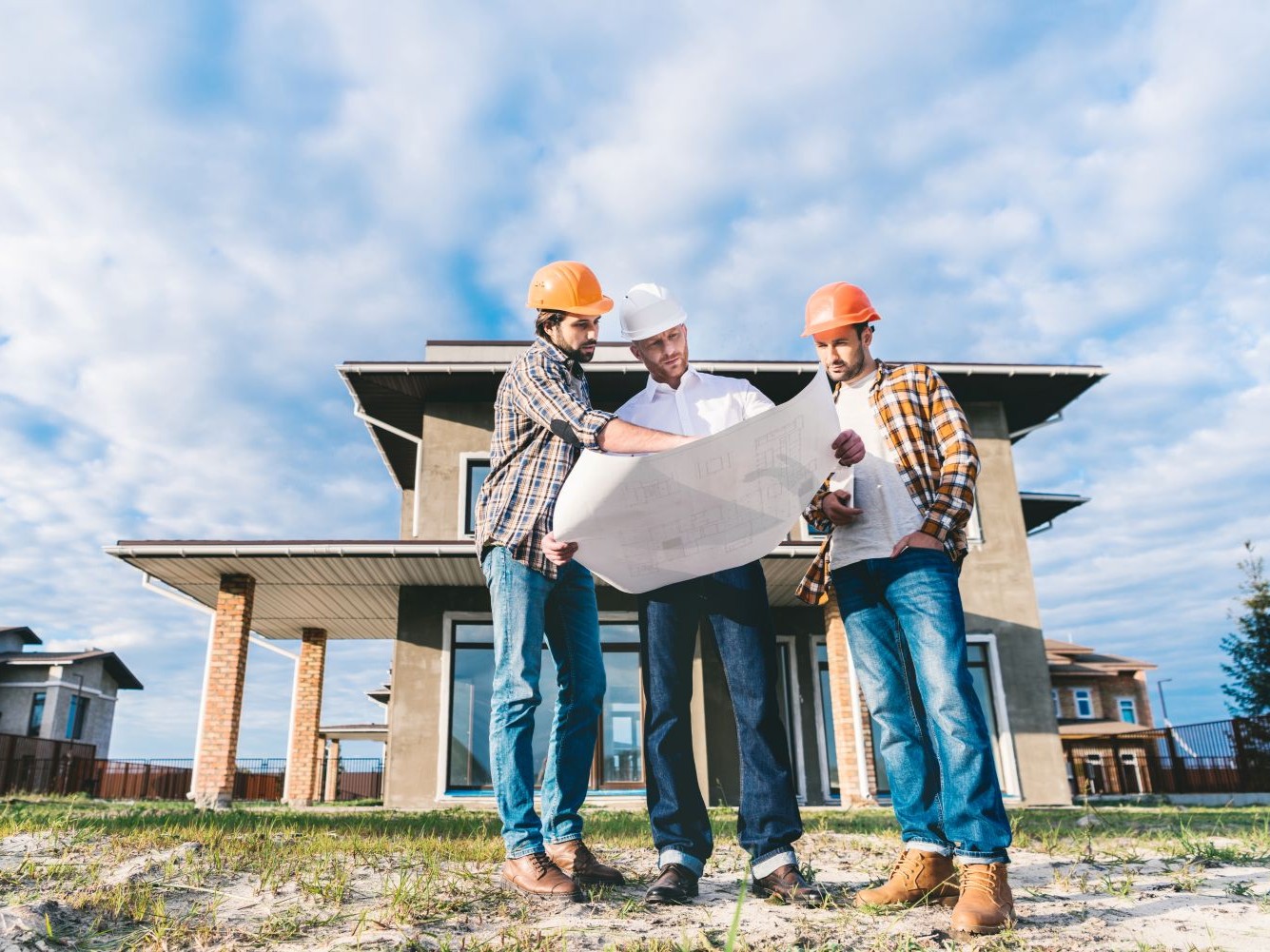 Three workers in hard hats reviewing a plan together in front of a two-storey house