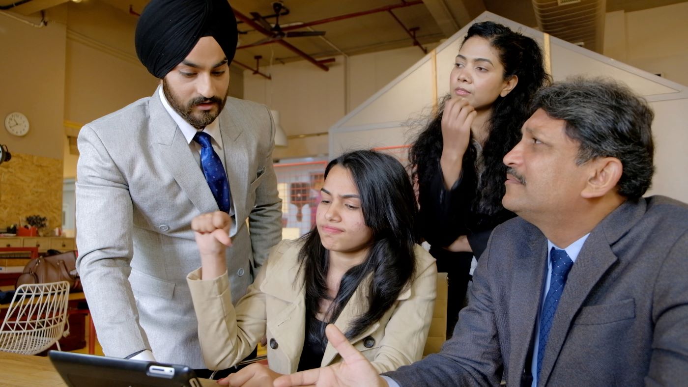 Colleagues in business dress gathered around a laptop at a meeting table, one man pointing at the screen