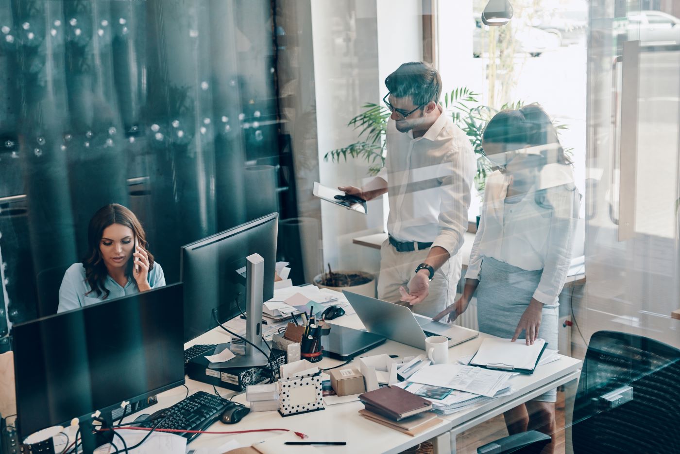 An open-plan office with several monitors, a woman on a phone call while colleagues talk behind her