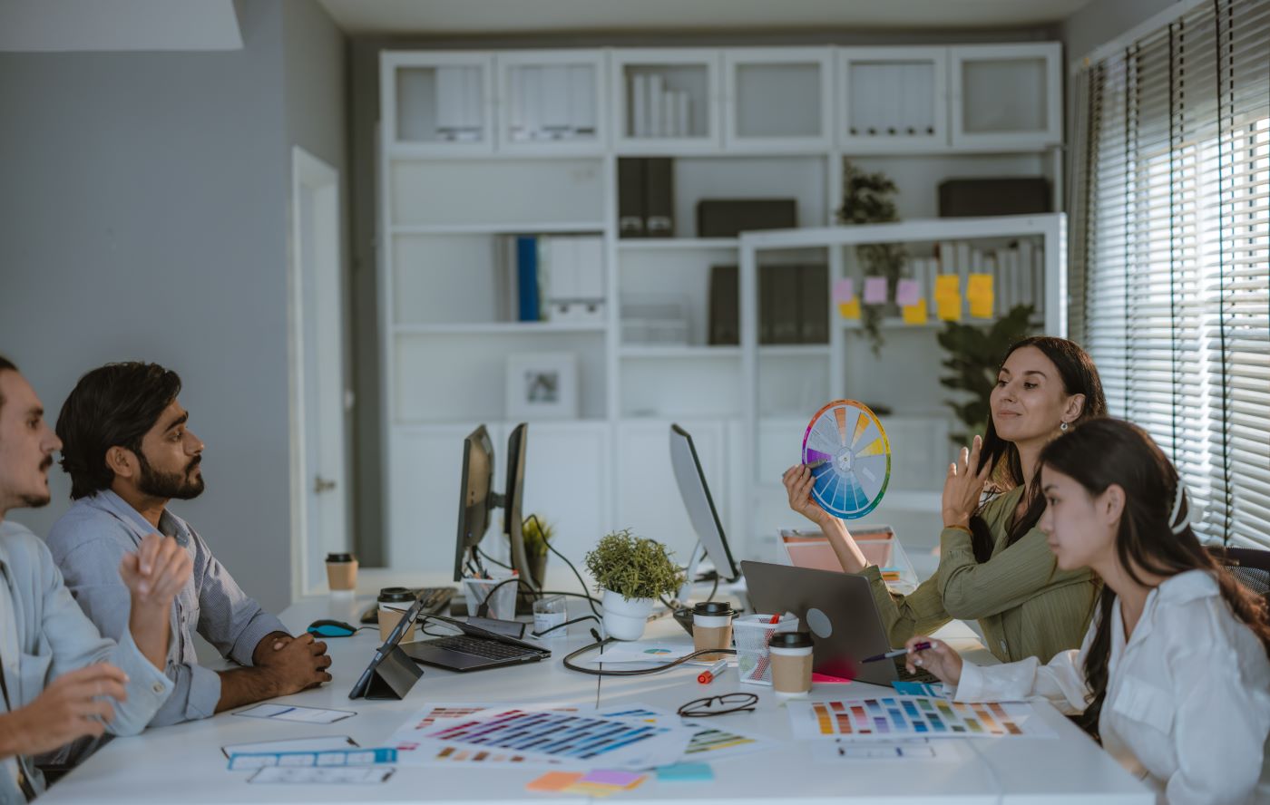 A design team at a shared desk covered in colour swatches, one woman holding up a colour wheel
