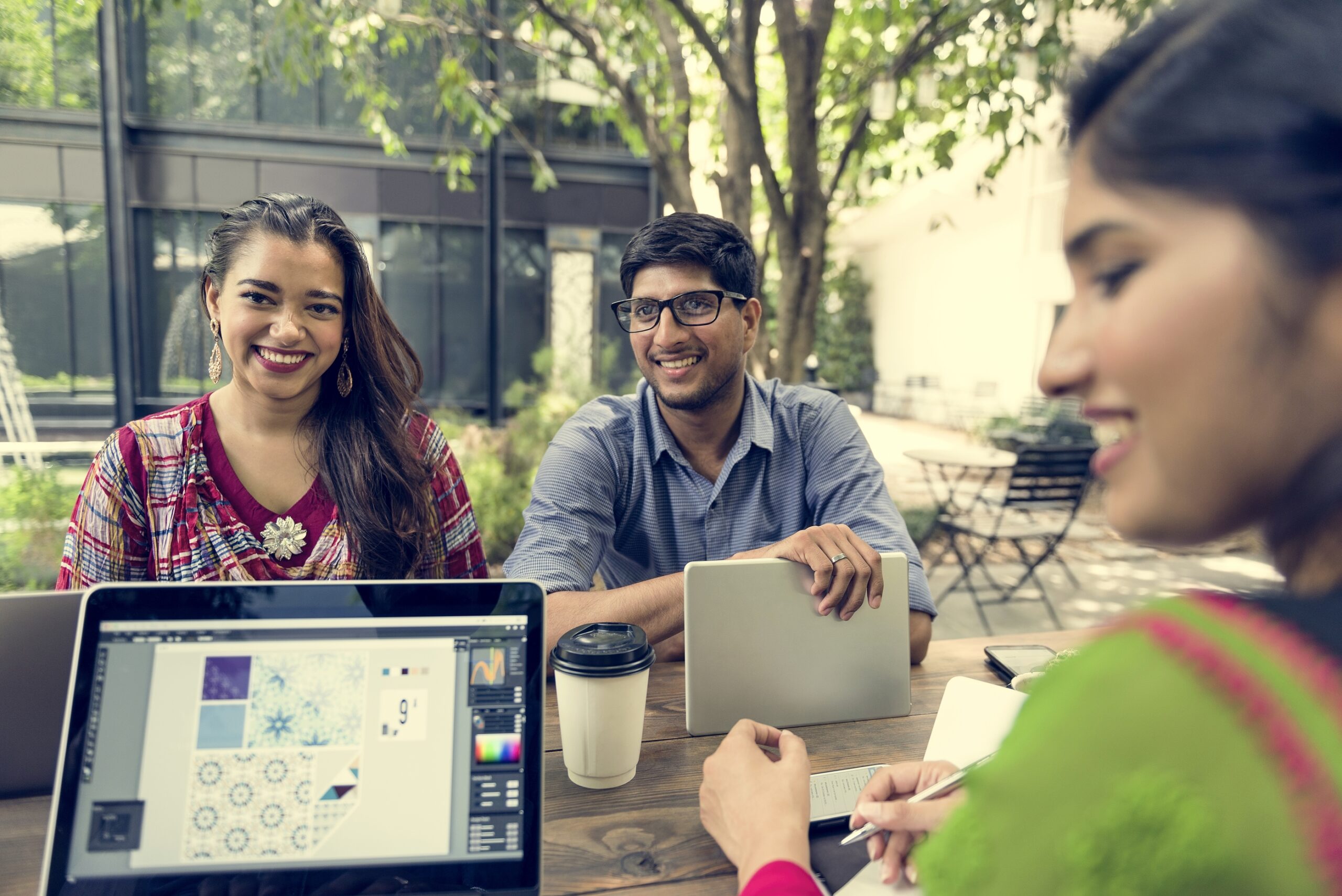 Three colleagues talking at an outdoor table with a laptop and coffee cups