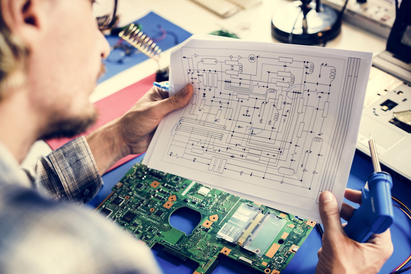 A technician holding a printed circuit diagram above a circuit board on a workbench, a soldering iron in hand