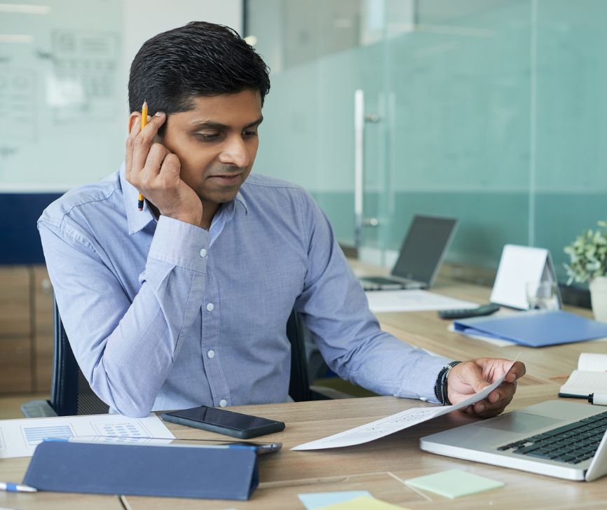 A man reviewing printed documents beside a laptop at an office desk