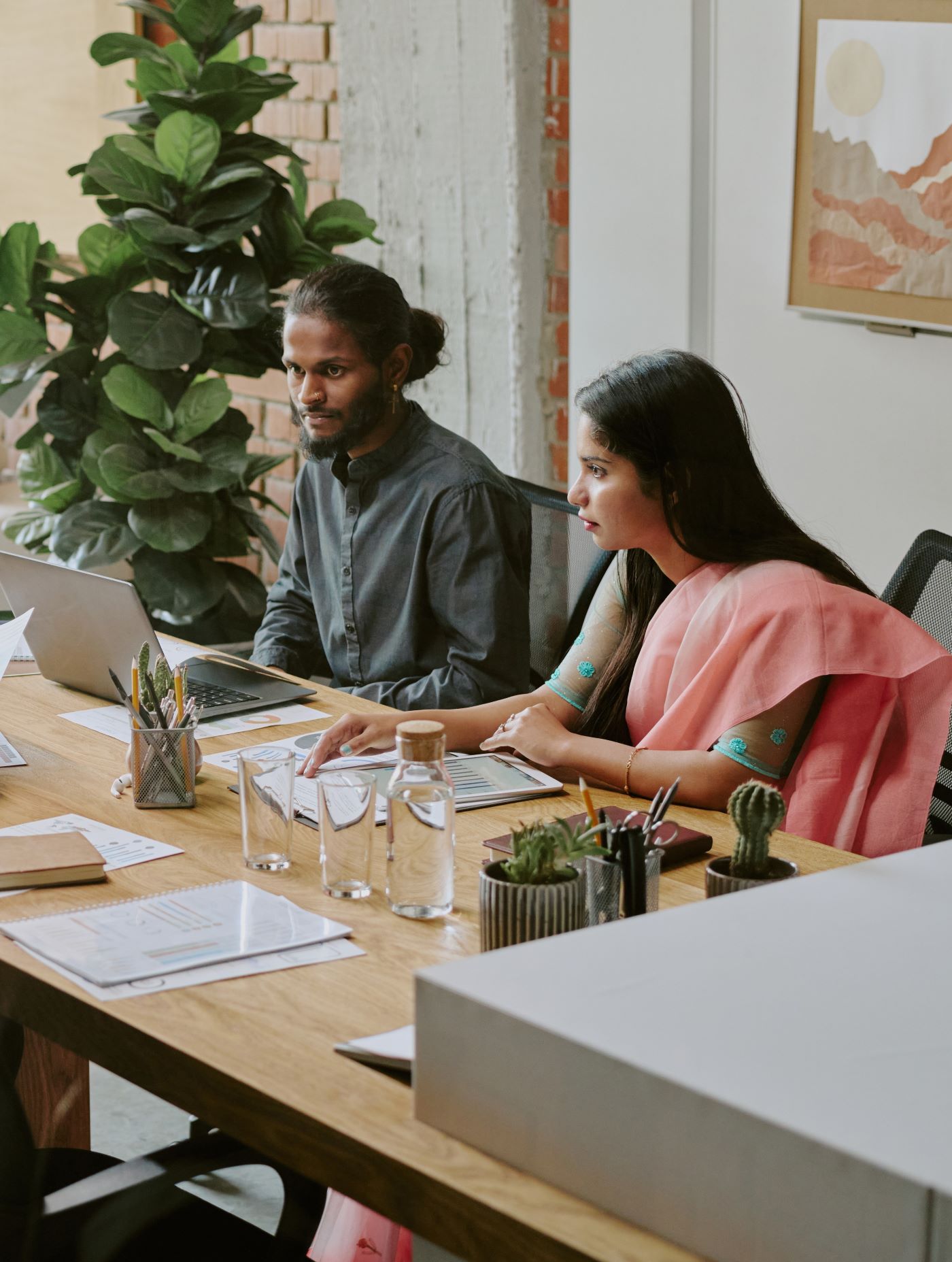 A man and a woman working at laptops on a shared office desk with plants