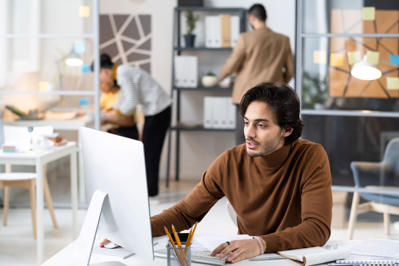 A developer working at a desktop computer in a bright open-plan office, colleagues at shelves behind him
