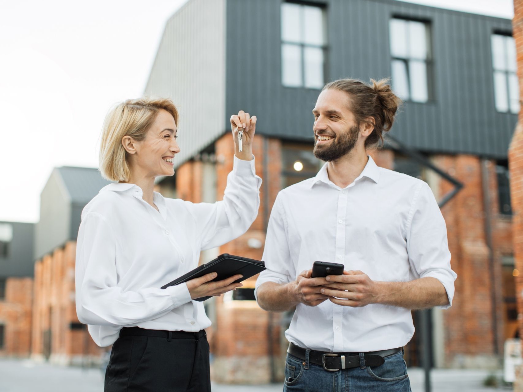 A woman holding up a set of keys to a smiling man outside a brick building, a tablet in her other hand