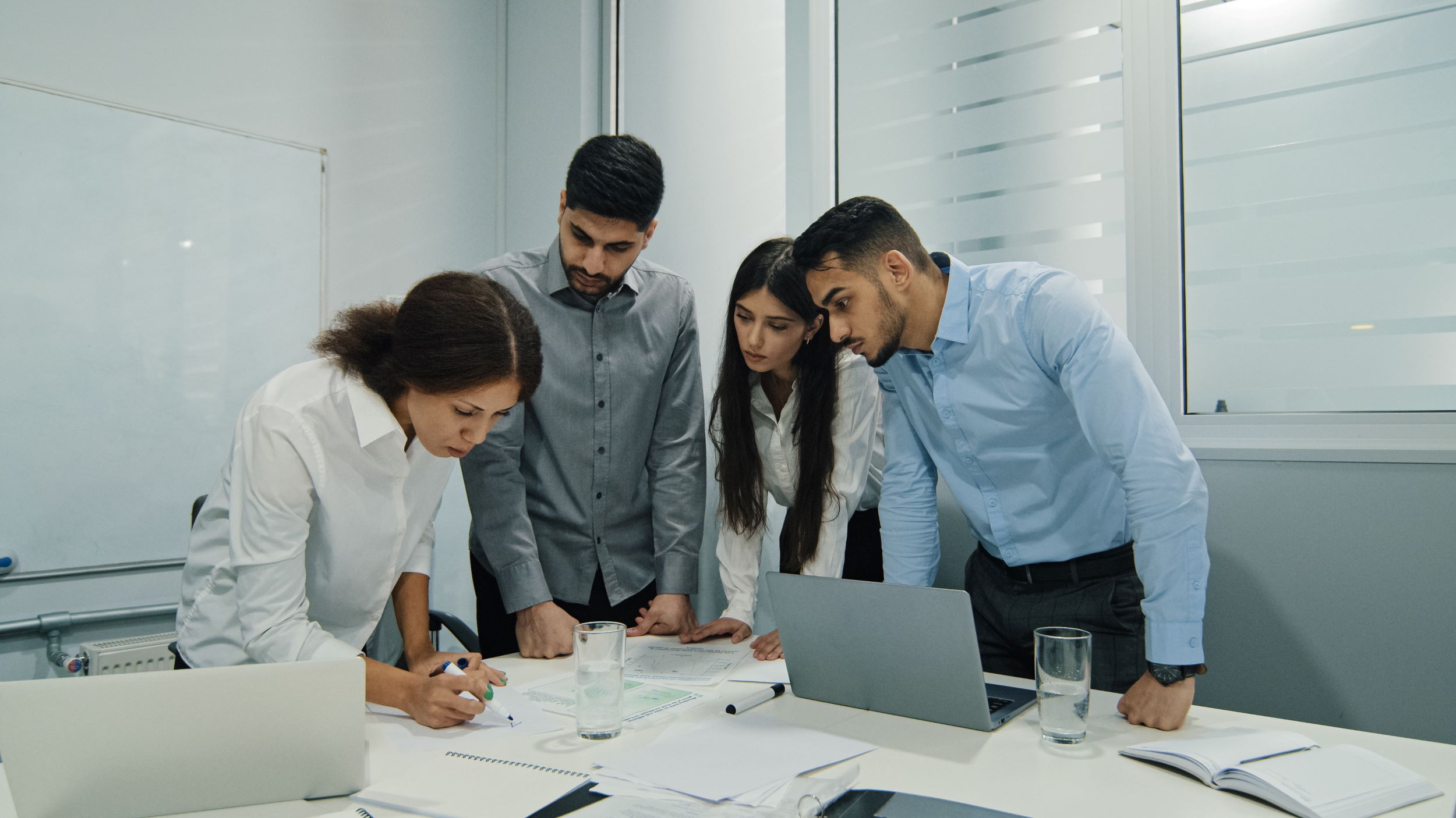 Four colleagues leaning over a meeting-room table to look at notes one of them is writing