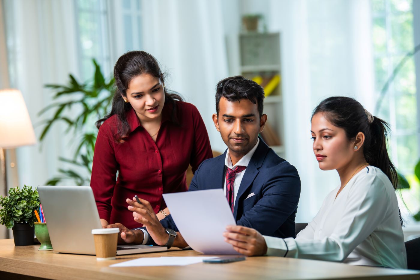 Three colleagues at a desk reviewing a printed document beside a laptop
