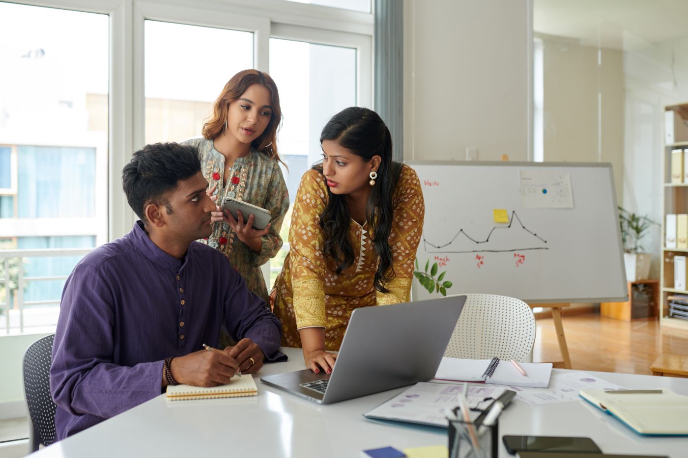 Three colleagues around a laptop at an office table, a whiteboard with a line chart behind them