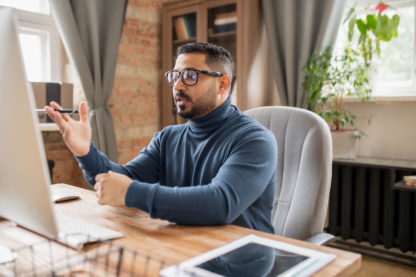 A man in a turtleneck and glasses gesturing at a desktop monitor in a brick-walled studio office