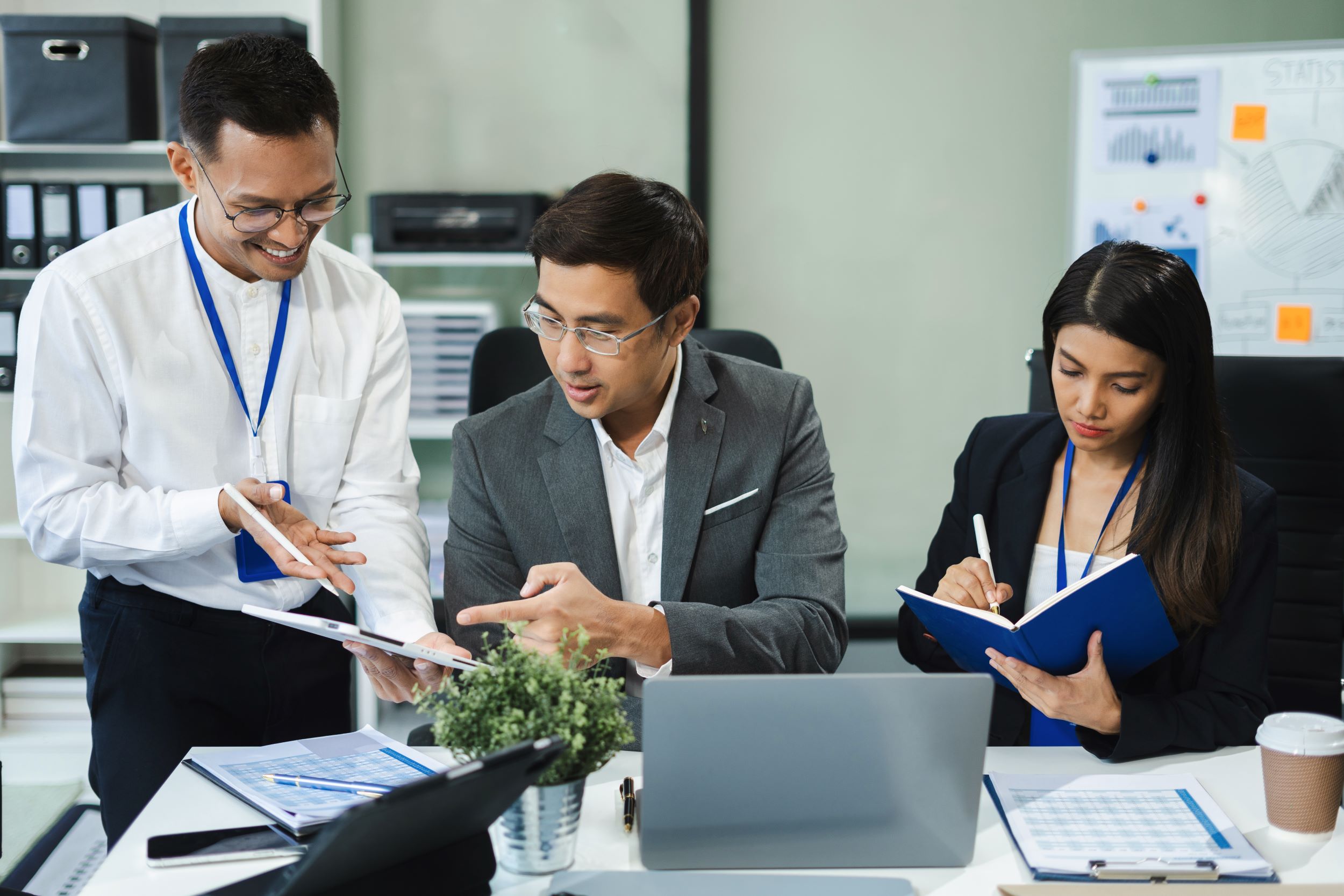 Colleagues with lanyards around a meeting table, one pointing at a tablet while another takes notes