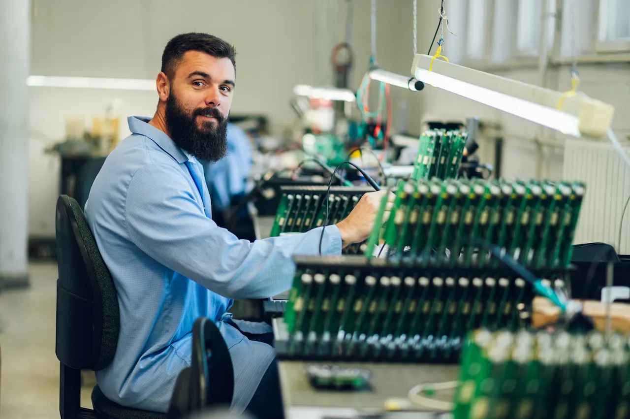 A technician seated at an electronics assembly bench lined with racks of circuit boards