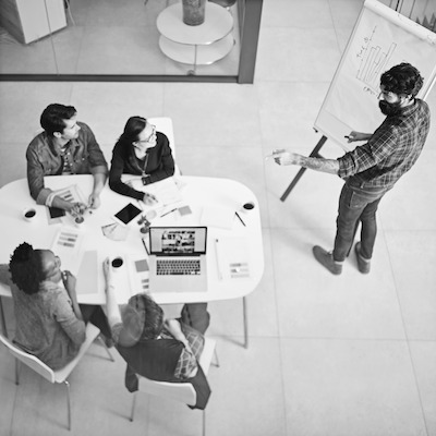 An overhead view of a team planning around a table while a colleague writes on a whiteboard