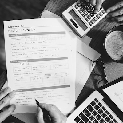 Hands completing a printed health insurance form beside a calculator and a laptop