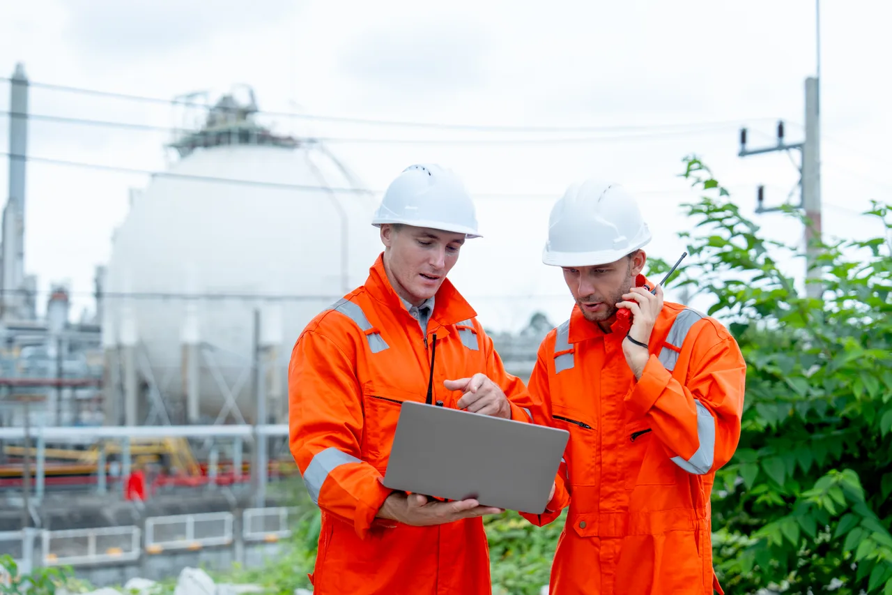 Two engineers in coveralls working on a laptop beside storage tanks