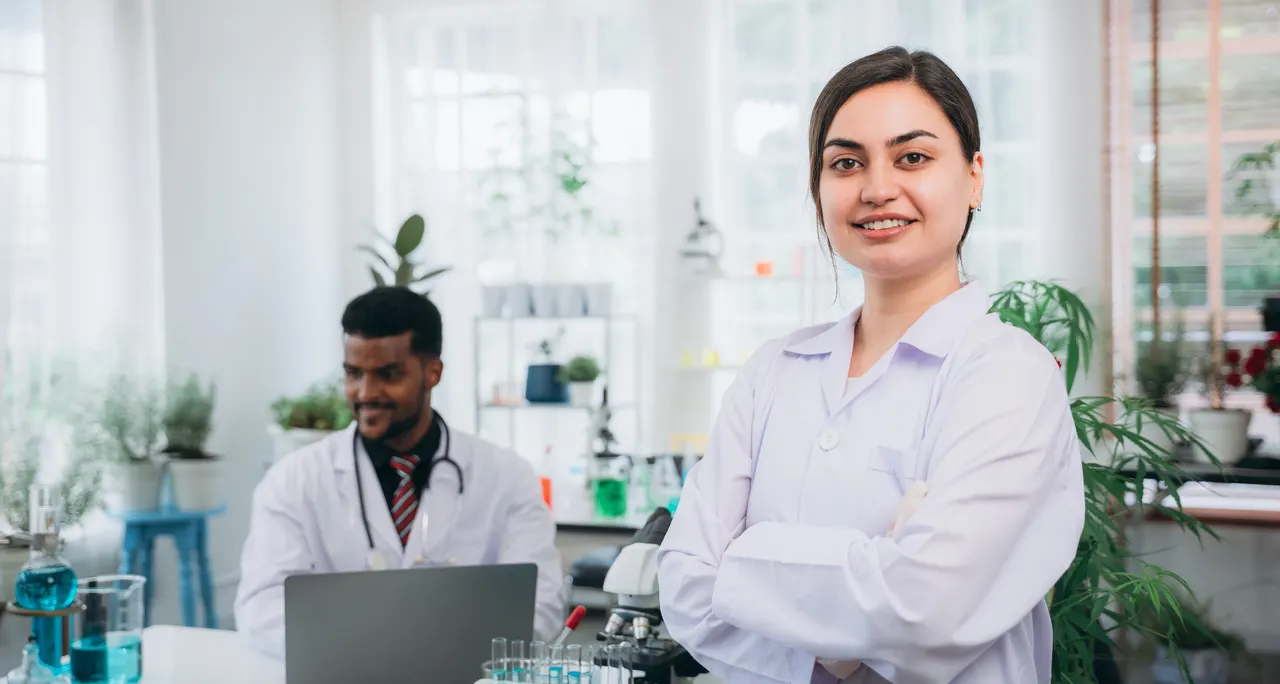 A laboratory analyst in a white coat with a colleague working at a laptop on the bench behind her