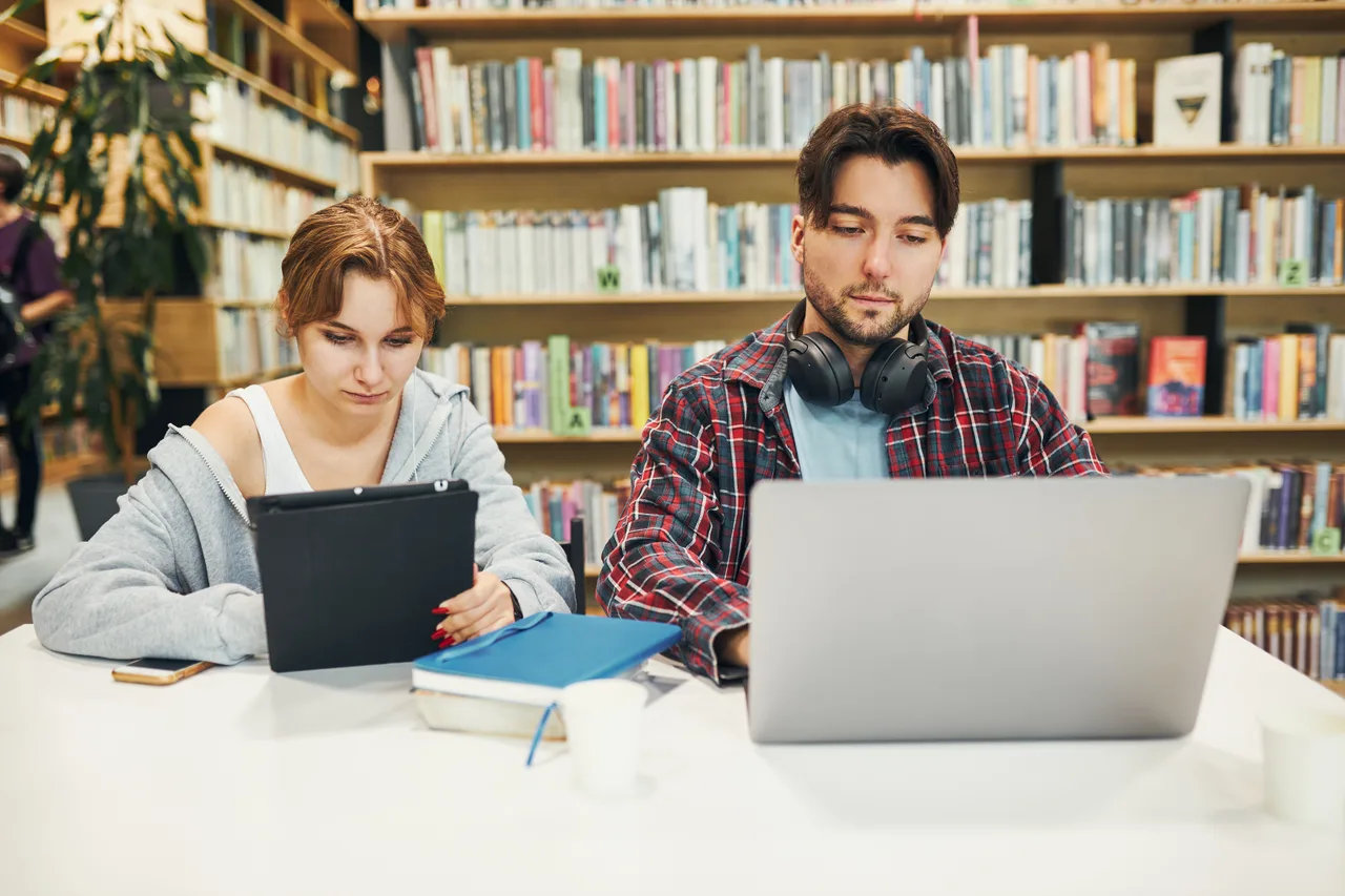 Two university students studying on laptops in a library