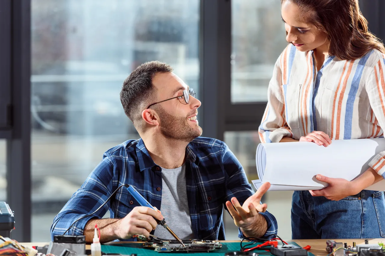 An engineer soldering a circuit board while a colleague reviews the plans